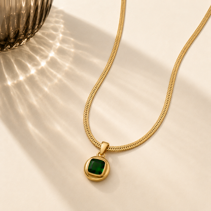 A gold chain necklace with a square green pendant set in a gold bezel, displayed on a warm cream surface with artistic light shadows.